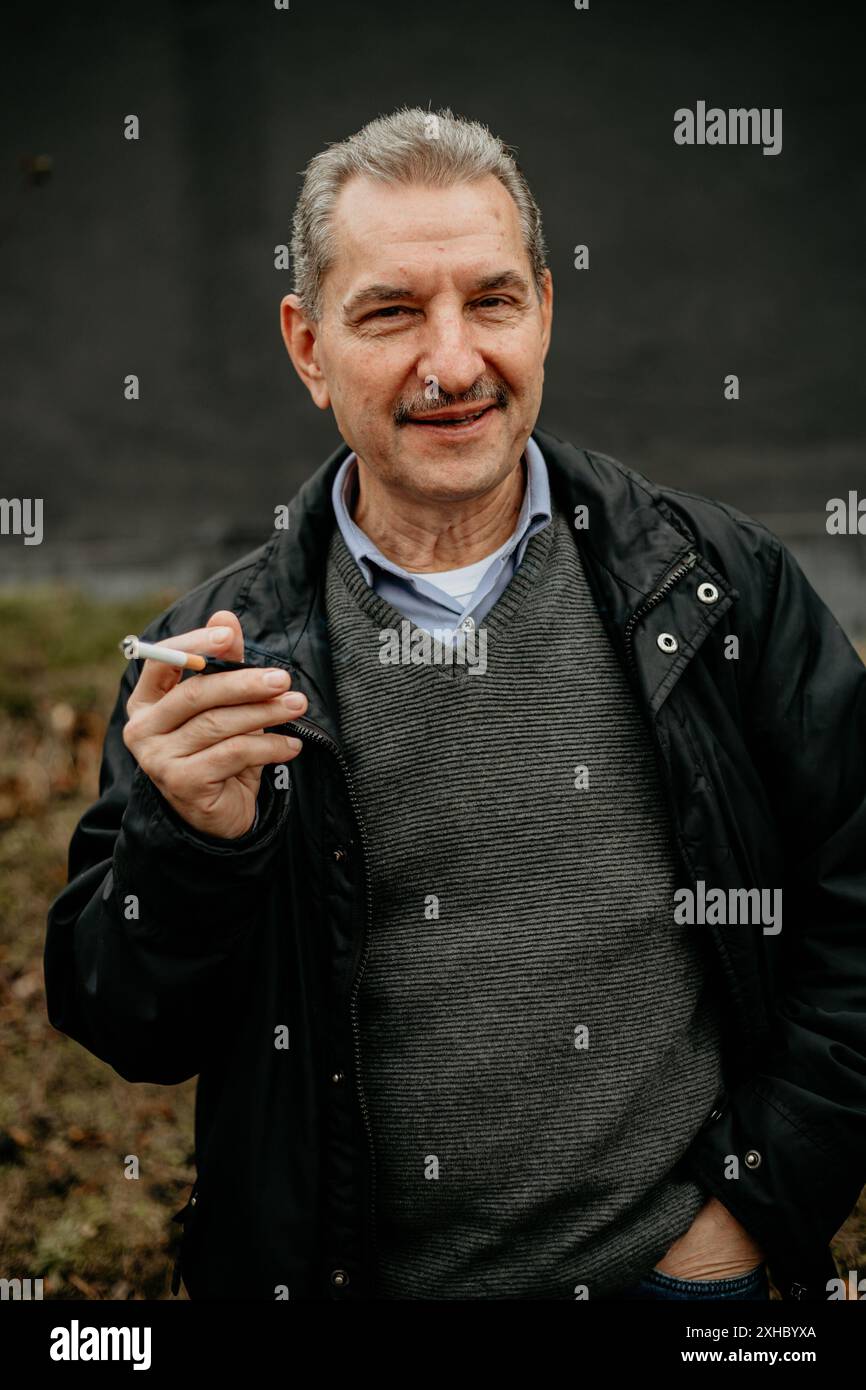 Ein älterer Mann, der eine schwarze Jacke und einen grauen Pullover trägt, steht draußen und raucht eine Zigarette. Er lächelt und schaut in die Kamera. Stockfoto