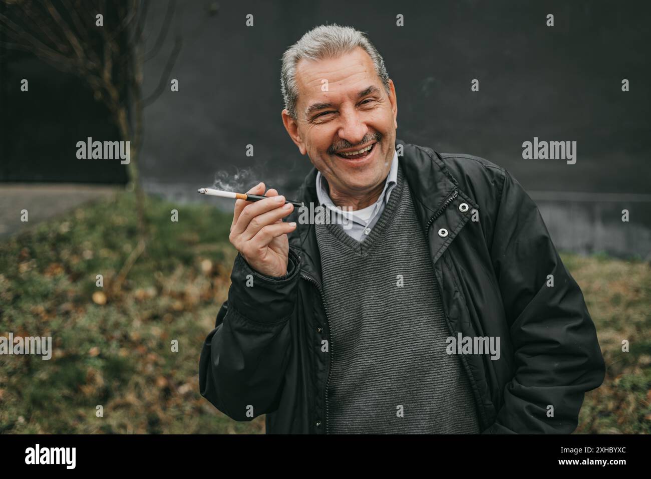 Ein Mann mit grauen Haaren, der eine schwarze Jacke und einen grauen Pullover trägt, raucht eine Zigarette. Er lächelt und schaut in die Kamera. Stockfoto