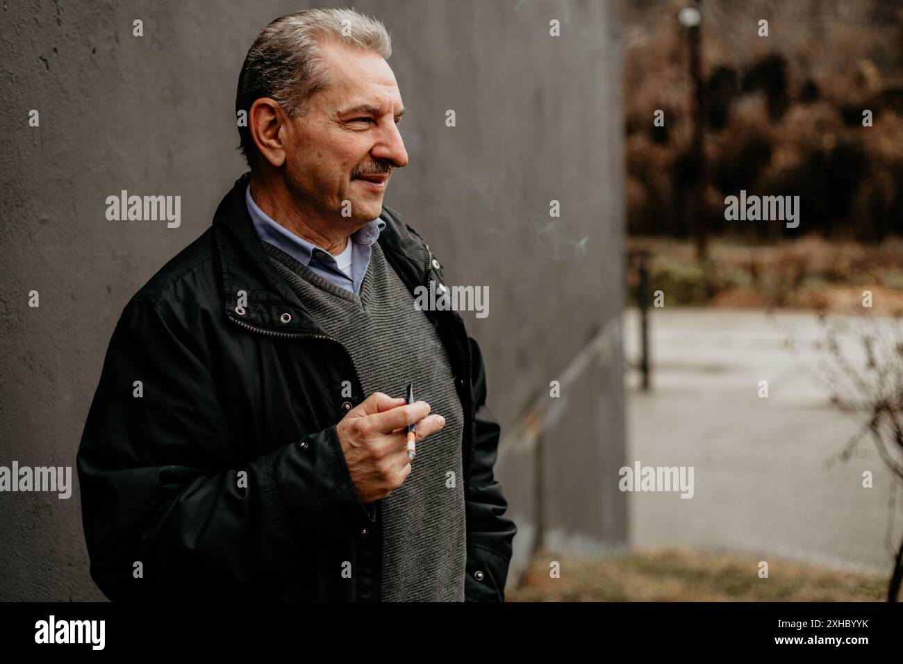 Ein Mann in einer schwarzen Jacke und einem grauen Pullover steht an einer grauen Wand und schaut zur Seite, während er eine Zigarette raucht. Stockfoto