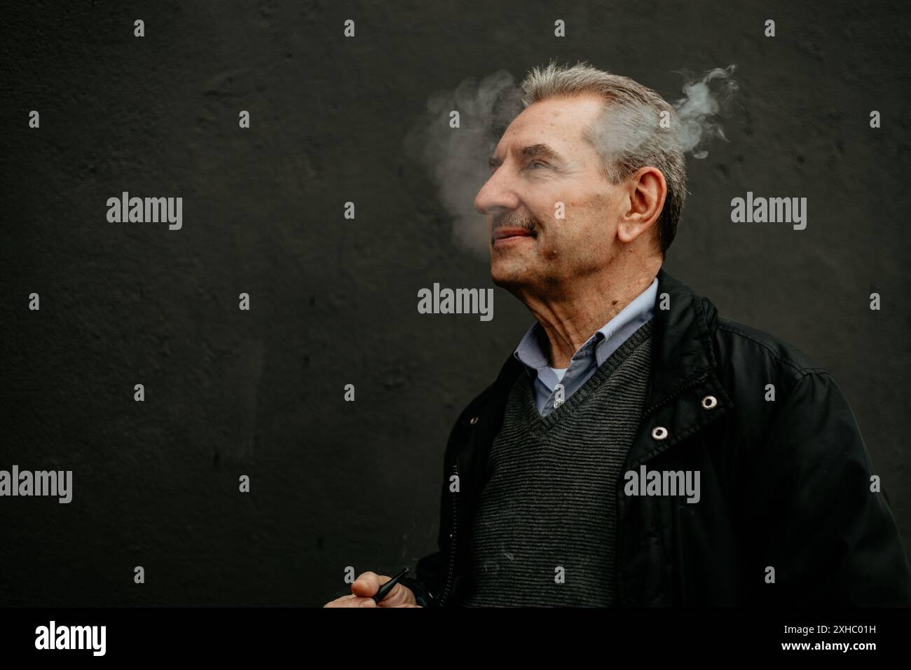 Ein älterer Mann mit grauen Haaren und Bart steht vor einer dunkelgrauen Wand und atmet einen Rauch aus. Stockfoto