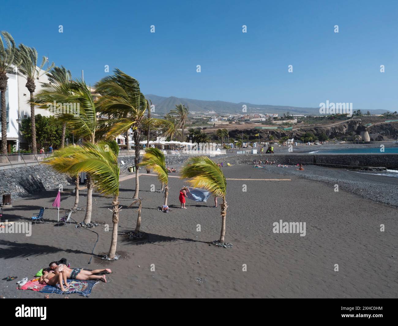 Playa de San Juan, Kanarische Insel, Teneriffa, Spanien, 1. Februar, 2024: schwarzer Sandstrand mit Sonnenbaden und kleinem Hafen. Playa de San Stockfoto