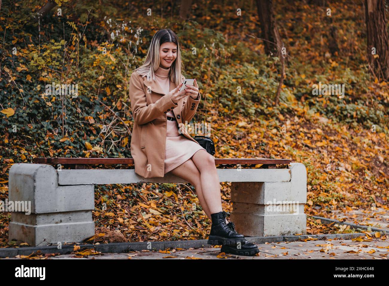 Eine Frau im braunen Mantel sitzt auf einer Bank im Park und benutzt ihr Telefon. Sie trägt ein pinkfarbenes Kleid und schwarze Stiefel. Der Boden ist mit abgefallenem Laub bedeckt Stockfoto