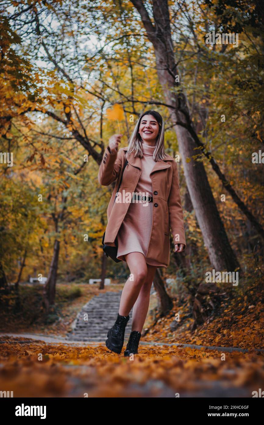 Eine Frau in braunem Mantel geht durch einen Herbstwald und lächelt, während sie ein gefallenes Blatt hält. Stockfoto