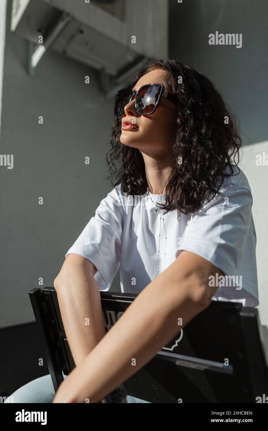 Cooles, schönes, modisches Mädchen-Model mit lockiger Frisur und trendiger Sonnenbrille in einem weißen T-Shirt sitzt bei Sonnenuntergang auf der Straße Stockfoto