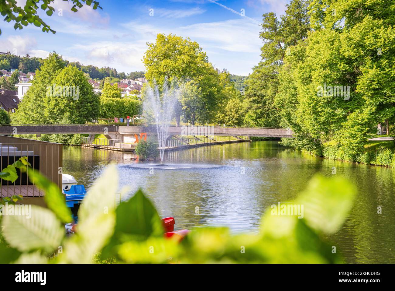 Ein Brunnen, der Wasser in einen Fluss sprüht, umgeben von Bäumen und einer Brücke, mit einer Stadt im Hintergrund, Nagold, Schwarzwald, Deutschland Stockfoto