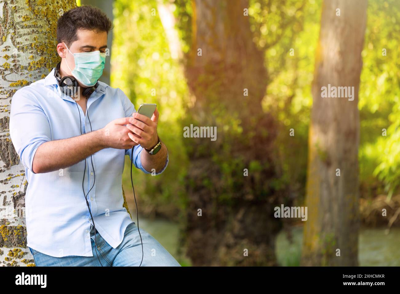 Junger Mann mit Schutzmaske, Musik im Freien im Park zu hören, während soziale Distanz beibehalten. Lebensstil während der Covid-19-Pandemie Stockfoto