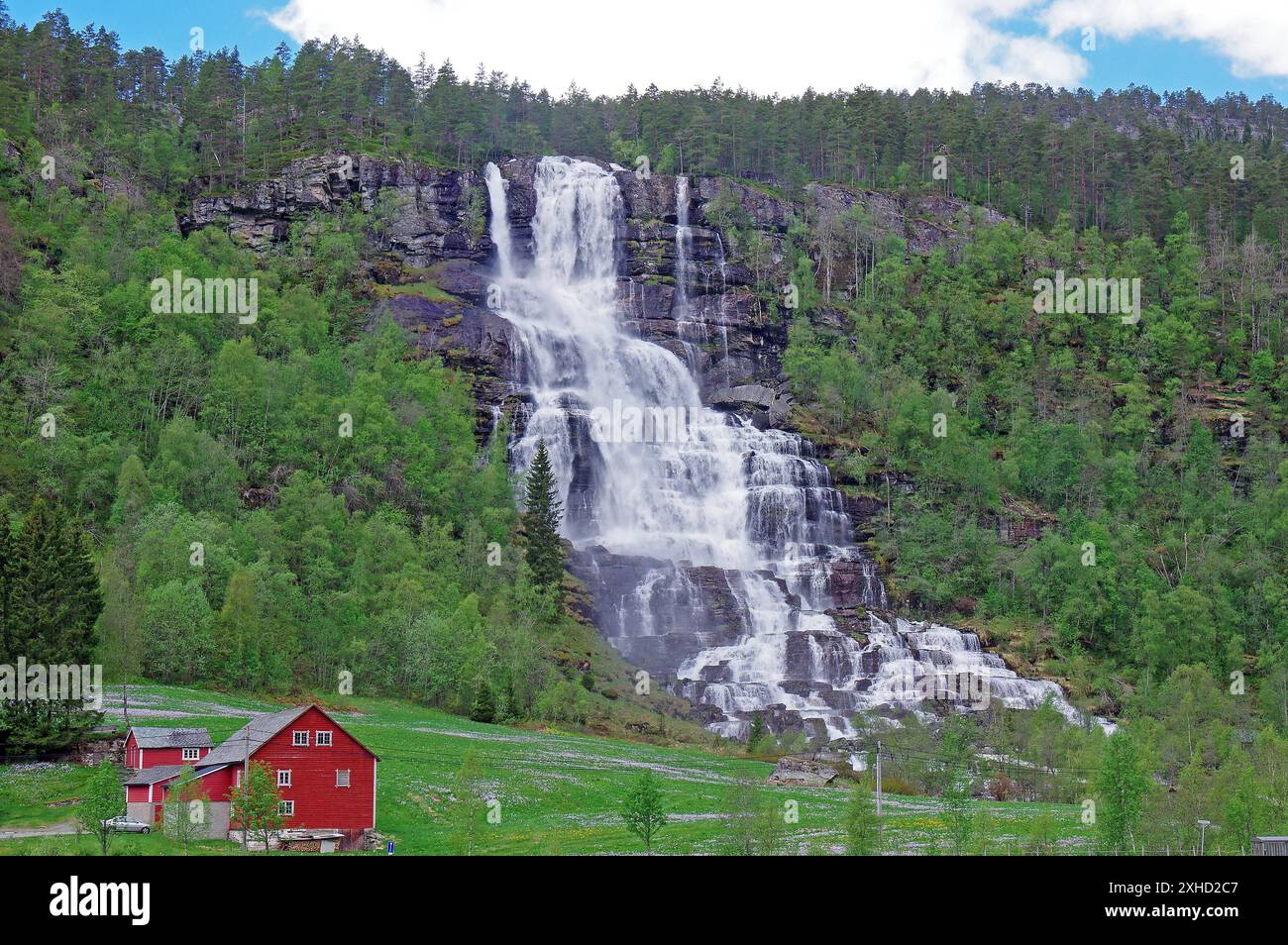 Ein großer Wasserfall in einer grünen Waldlandschaft mit roten Häusern und Bergen im Hintergrund, Tvindefossen, Norwegen Stockfoto