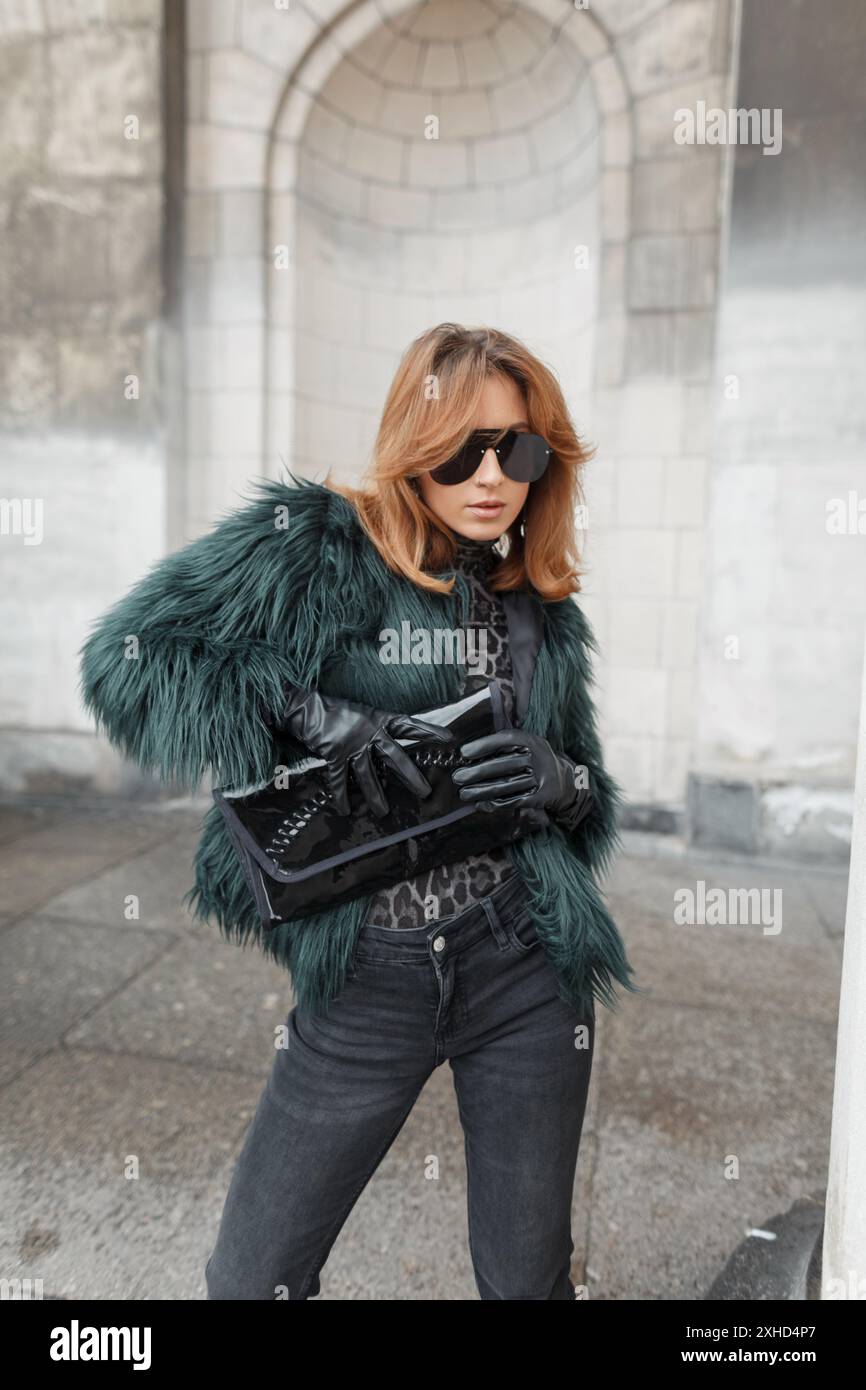 Mode schöne urbane Vogue Model Frau mit Sonnenbrille in einer stilvollen zotteligen Jacke mit einer Handtasche, die in der Stadt in der Nähe des Gebäudes posiert Stockfoto