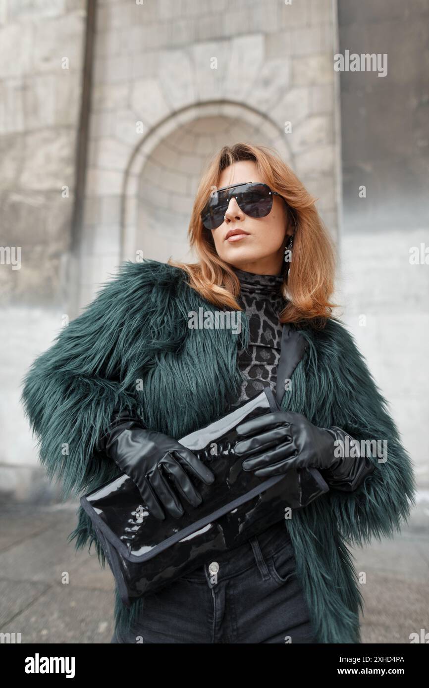 Cooles, stilvolles, glamouröses Damenmodell mit cooler Sonnenbrille in modischer Kleidung mit zotteliger Jacke mit Handschuhen und Handtasche in der Stadt Stockfoto