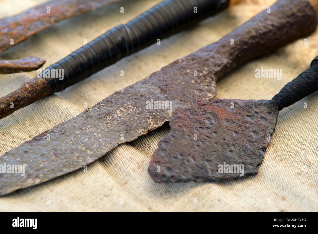 Ein rostiges Messer ist neben einem rostigen Speer. Das Messer ist alt und hat ein schwarzes Band. Die beiden Objekte befinden sich auf einem weißen Tuch Stockfoto