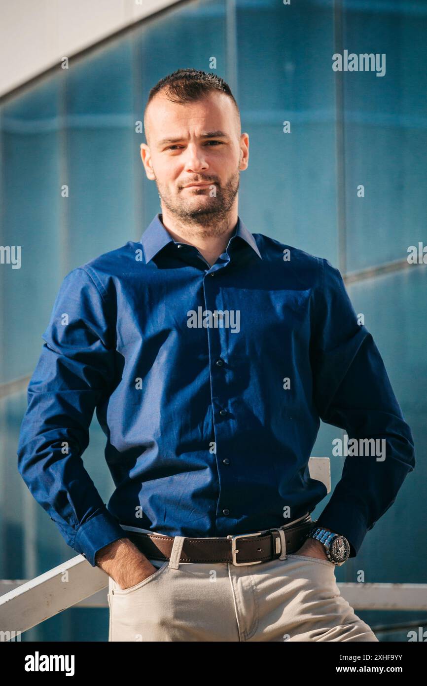 Ein Mann in einem blauen Hemd mit ernstem Ausdruck steht vor einem modernen Glasgebäude. Stockfoto