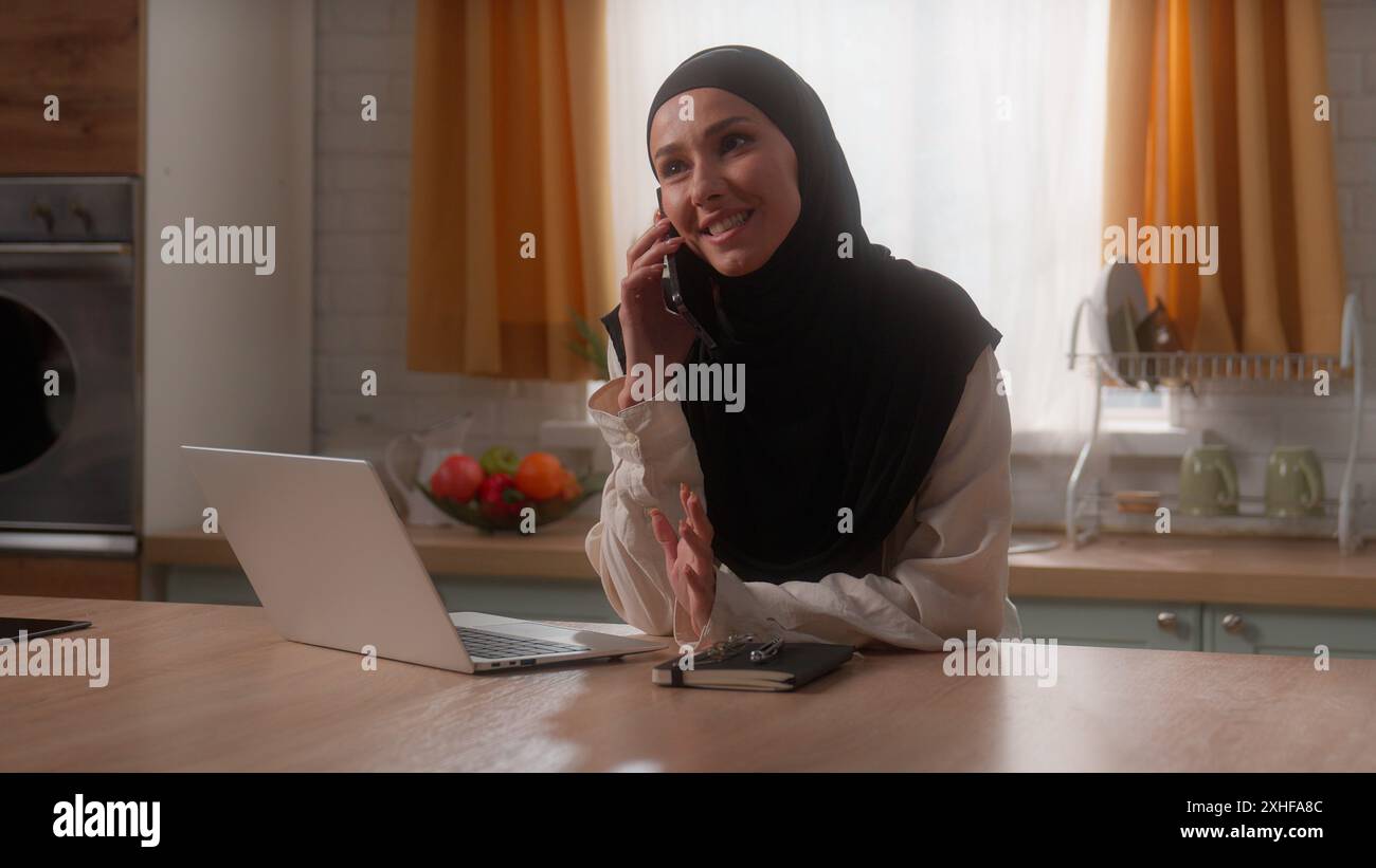 Glückliches lächelndes islamisches Mädchen muslimische Frau im Hijab Dame indische weibliche Hausfrau Klientin bestellen Essenslieferung in der Küche mit Laptop sprechendem Handy Stockfoto