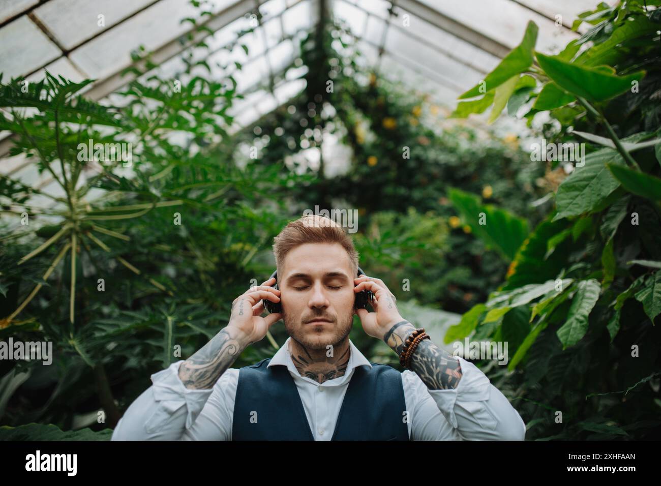 Gutaussehender Mann mit Tätowierung, der Meditationsmusik im botanischen Garten zuhört, inmitten von üppig grünem Laub. Er kümmert sich um seine psychische Gesundheit Stockfoto