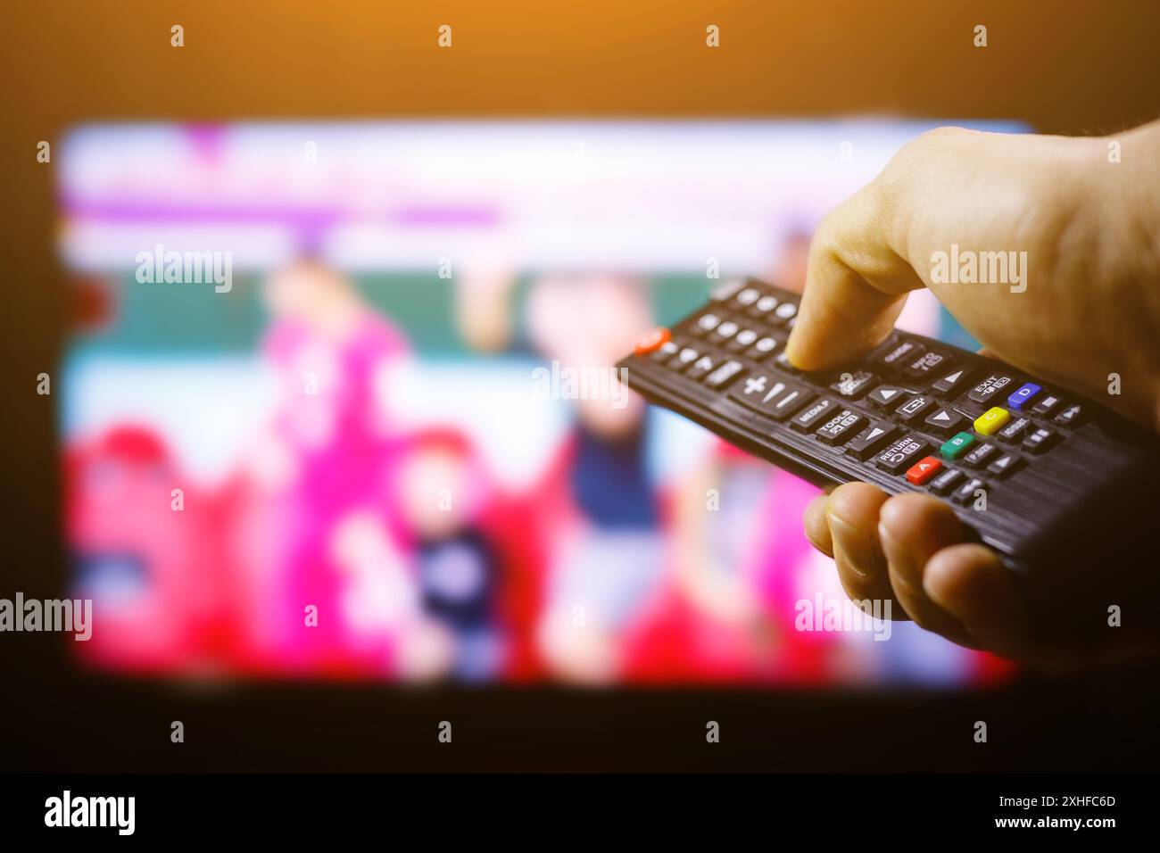 Eine Hand drückt eine Taste auf einer schwarzen Fernbedienung, wobei ein unscharfer Fernsehbildschirm im Hintergrund angezeigt wird. Stockfoto