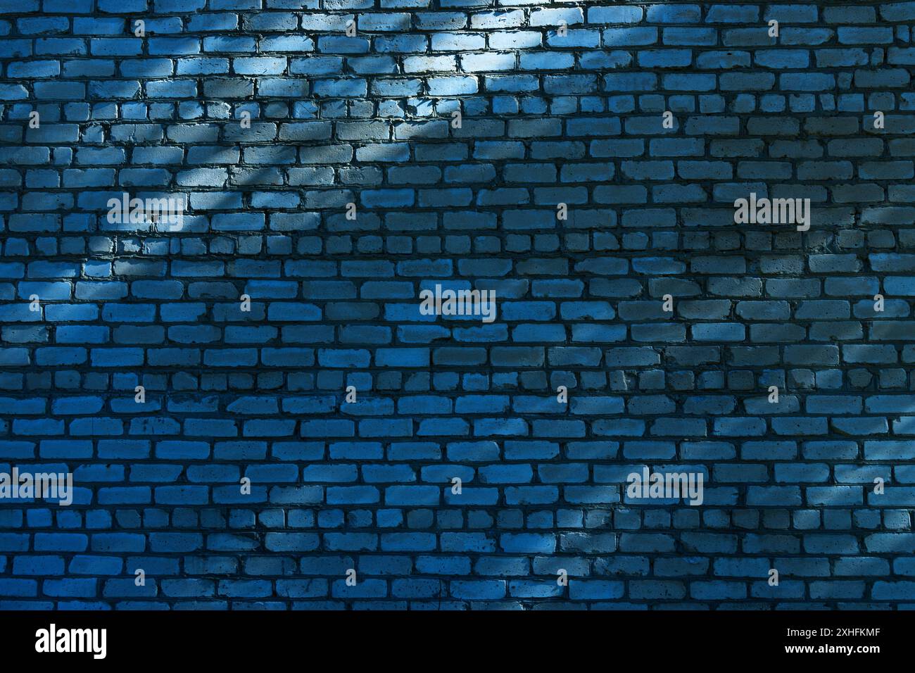 Eine blaue Ziegelwand mit diagonalem Sonnenlicht wirft Schatten über die Oberfläche. Das Licht ist weich und die Schatten sind subtil, wodurch eine visuell interessante Textur entsteht. Stockfoto