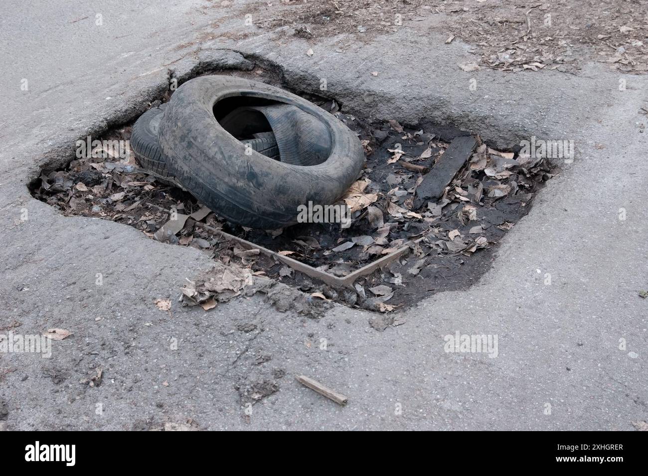 Alte, abgerissene Reifen liegen in einem Loch in der Asphaltstraße, mit den Überresten eines Sturmabflusses am Boden des Lochs Stockfoto