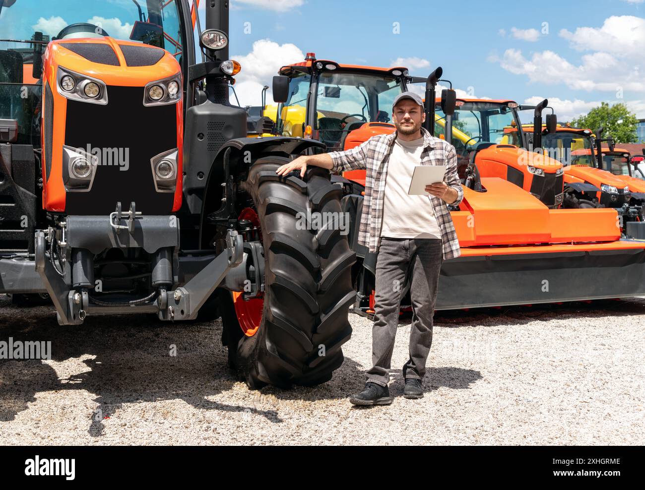 Kaukasischer Erwachsener mit digitalem Tablet in der Hand steht neben einem Bau- oder landwirtschaftlichen Traktor bei einem Maschinenhändler. AG-Gerätehändler oder -Zulieferer Stockfoto