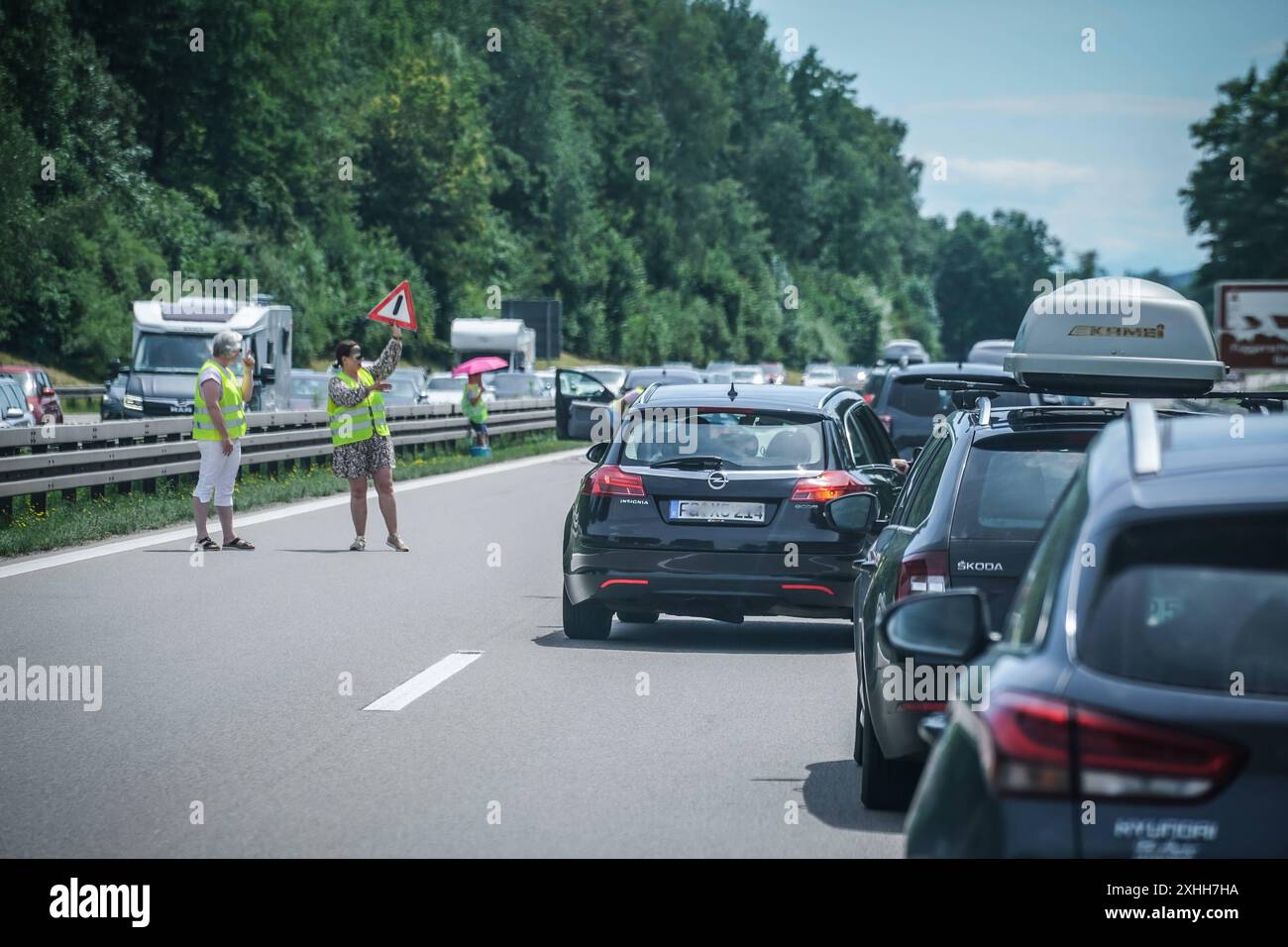 Szene auf einer Autobahn bei einem Verkehrsstau. Im Vordergrund sieht man mehrere Fahrzeuge, darunter einen Opel Insignia und einen Å koda, die auf der rechten Fahrspur stehen. Auf den Dächern der Fahrzeuge befinden sich Fahrradträger mit Fahrrädern sowie eine Dachbox. Zwei Personen in gelben Warnwesten stehen am Fahrbahnrand und halten ein Verkehrsschild hoch, um die Verkehrsteilnehmer auf eine Gefahrensituation hinzuweisen. *** Szene auf einer Autobahn während eines Staus im Vordergrund können Sie mehrere Fahrzeuge sehen, darunter ein Opel-Abzeichen und ein Å-Koda, die auf der rechten Spur auf der geparkt sind Stockfoto