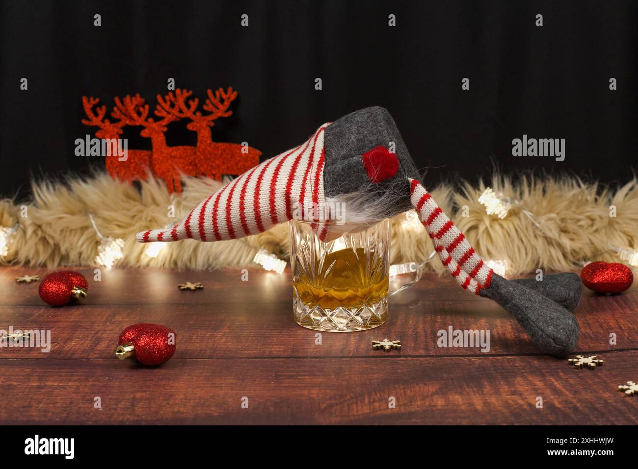 Ein betrunkener Weihnachtszwerg mit der Nase in einem Glas Whiskey auf einer Party. Im Hintergrund Rentiere, Herzen, Sterne und Schneeflocken geformte Feenlichter Stockfoto
