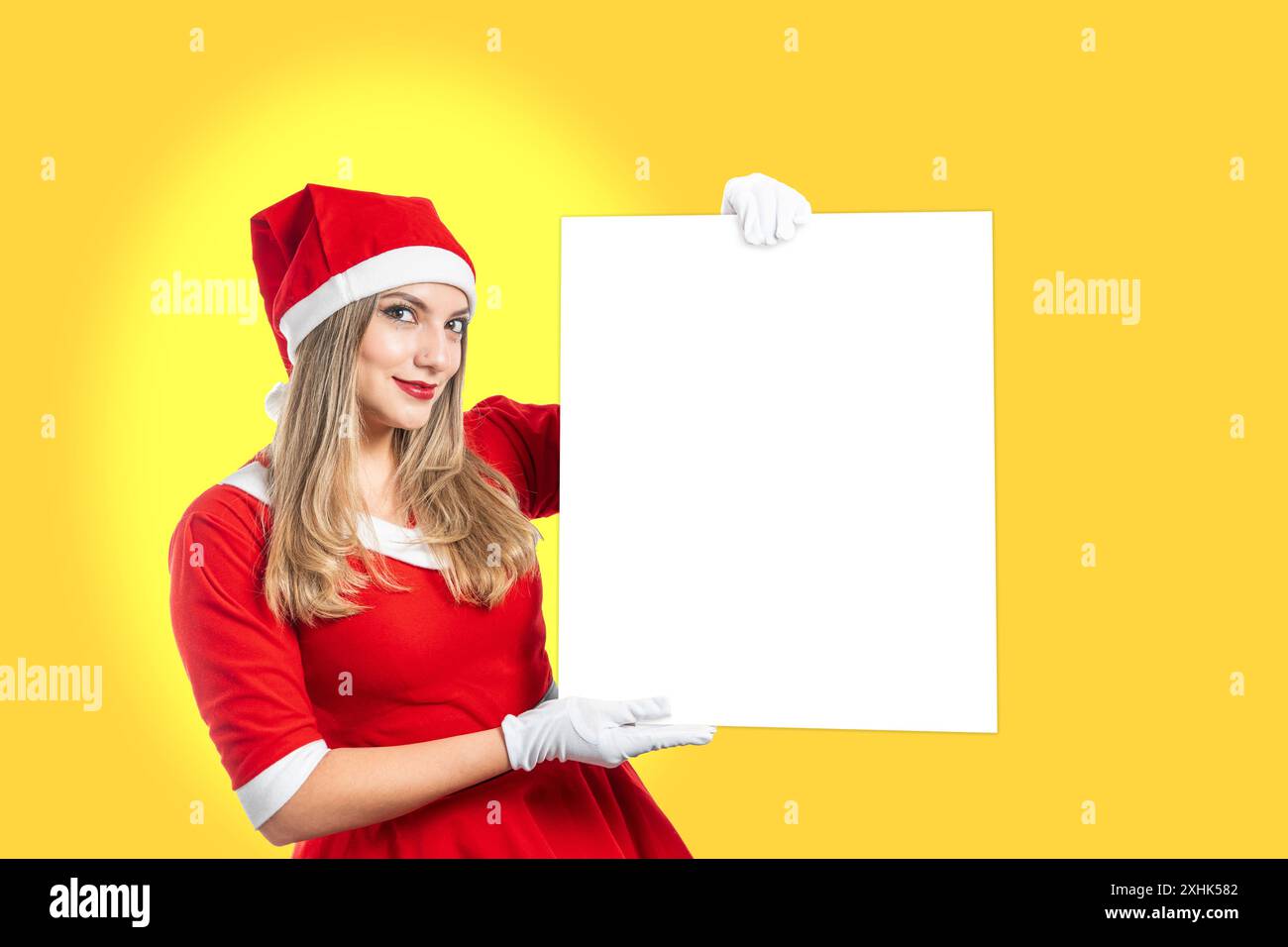 Eine junge Frau, die als Weihnachtsmann gekleidet ist, lächelt in die Kamera, die ein Schild mit Kopierraum auf gelbem Hintergrund zeigt. Horizontale Weihnachtsfotografie Stockfoto