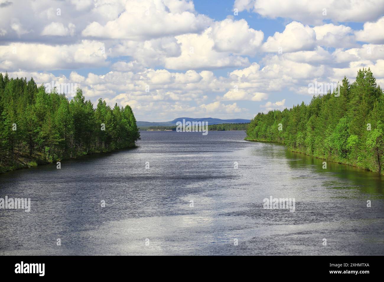 Der Fluss wird von üppigen grünen Wäldern unter einem Himmel mit weißen Wolken und fernen Bergen in Nordschweden flankiert. Stockfoto