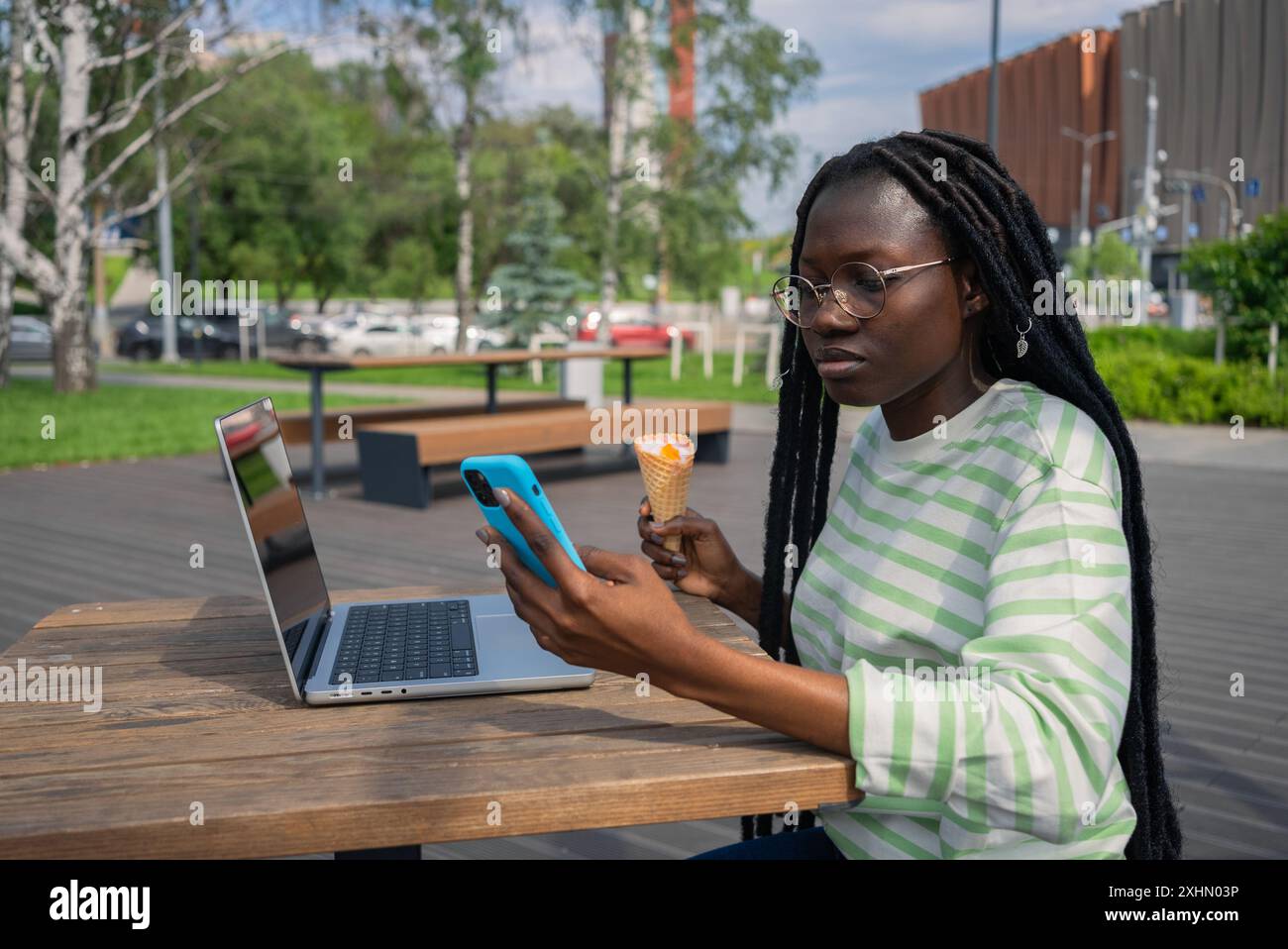Schwarze Frau genießt Eis am Tisch im Straßencafé. Junge Freelancerin macht Pause von der Arbeit am Laptop und genießt coole Desserts, die Nachrichten auf dem Smartphone lesen. Das Konzept der Vereinbarkeit von Beruf und Privatleben Stockfoto