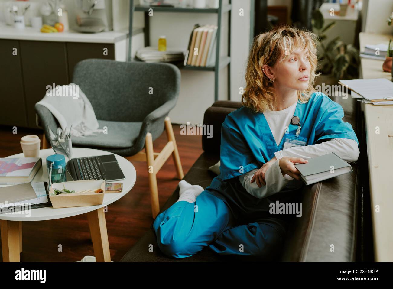 Gut aussehende kaukasische Krankenschwester, die auf einer schwarzen Ledercouch sitzt und ein graues Buch hält, während sie aus dem Fenster blickt und die Aussicht genießt Stockfoto