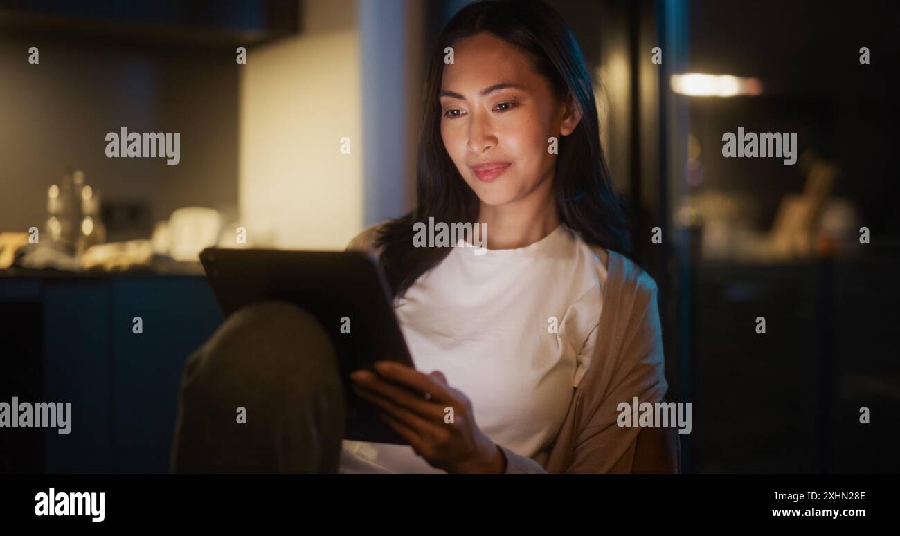 Schöne authentische asiatische Frau, die an einem Tisch in der gemütlichen Küche sitzt und nachts ein digitales Tablet verwendet. Frauen lächeln und chatten in sozialen Medien, machen Online-Shopping, surfen im Internet Stockfoto