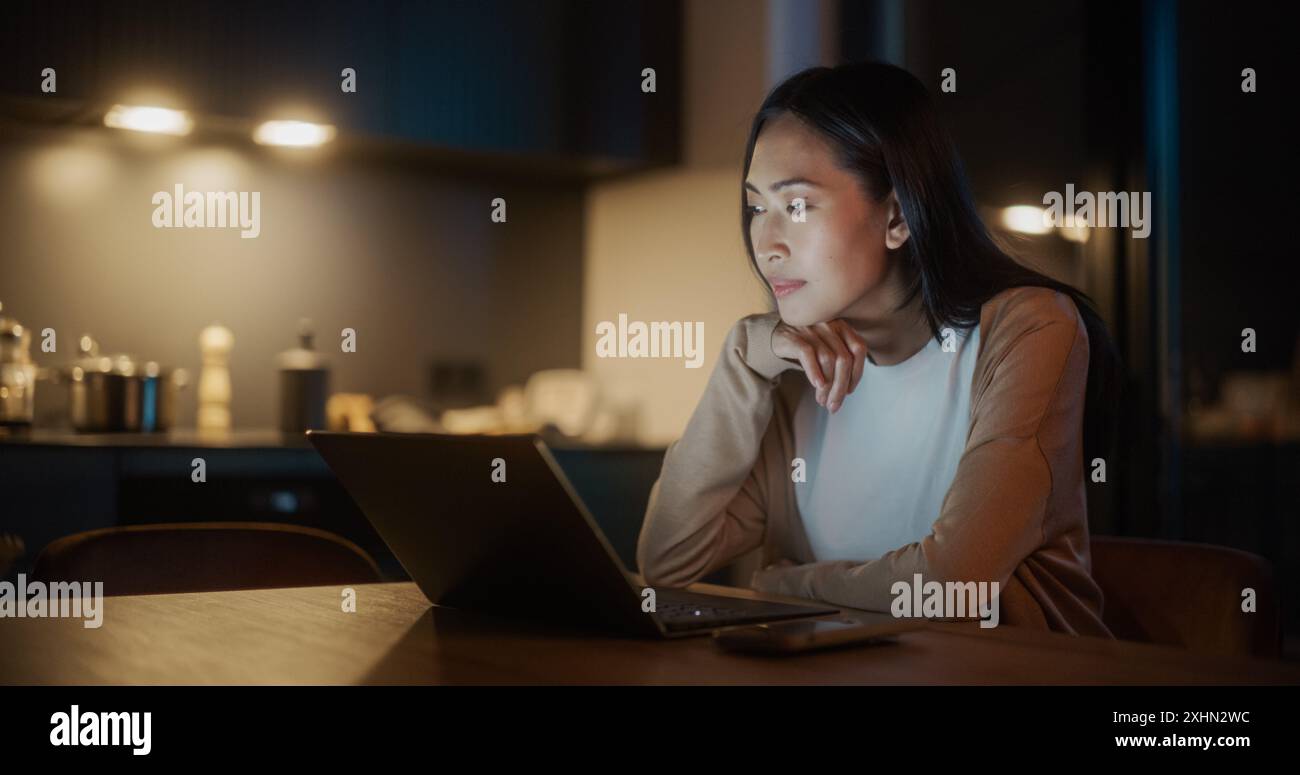 Schöne authentische asiatische Frau, die an einem Tisch in stilvoller Küche sitzt und nachts einen Laptop benutzt. Frauen lächeln und chatten in sozialen Medien, machen Online-Shopping, sehen sich die Nachrichten an. Stockfoto
