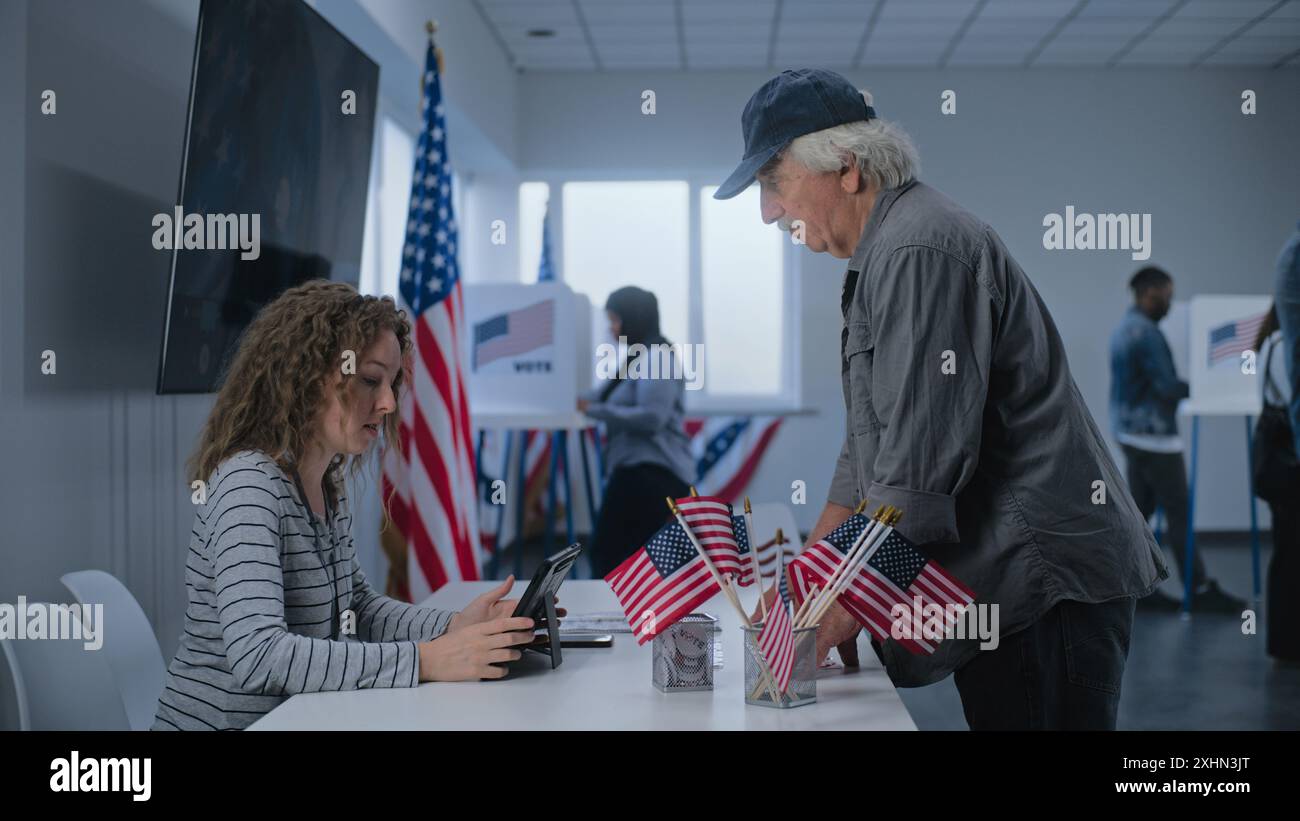 Kaukasischer reifer US-Bürger steht in der Nähe der Registrierungstafel am Wahlschalter. Die weibliche Wahlverantwortliche spricht mit dem Mann, benutzt einen Tablet-Computer und gibt Stimmzettel für die Wahl. Wahltag in den USA. Stockfoto