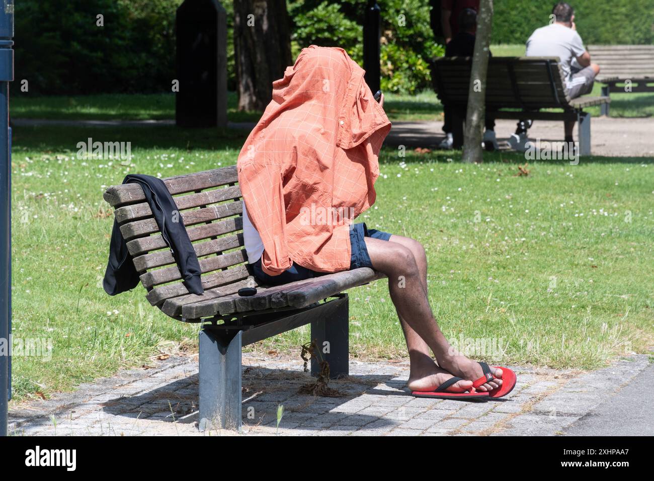 Mann, der auf einer Parkbank sitzt und den Kopf mit einem Hemd bedeckt hat, England, Großbritannien. Konzept: Obdachlose, Obdachlosigkeit, illegale Einwanderer, Einwanderer Stockfoto