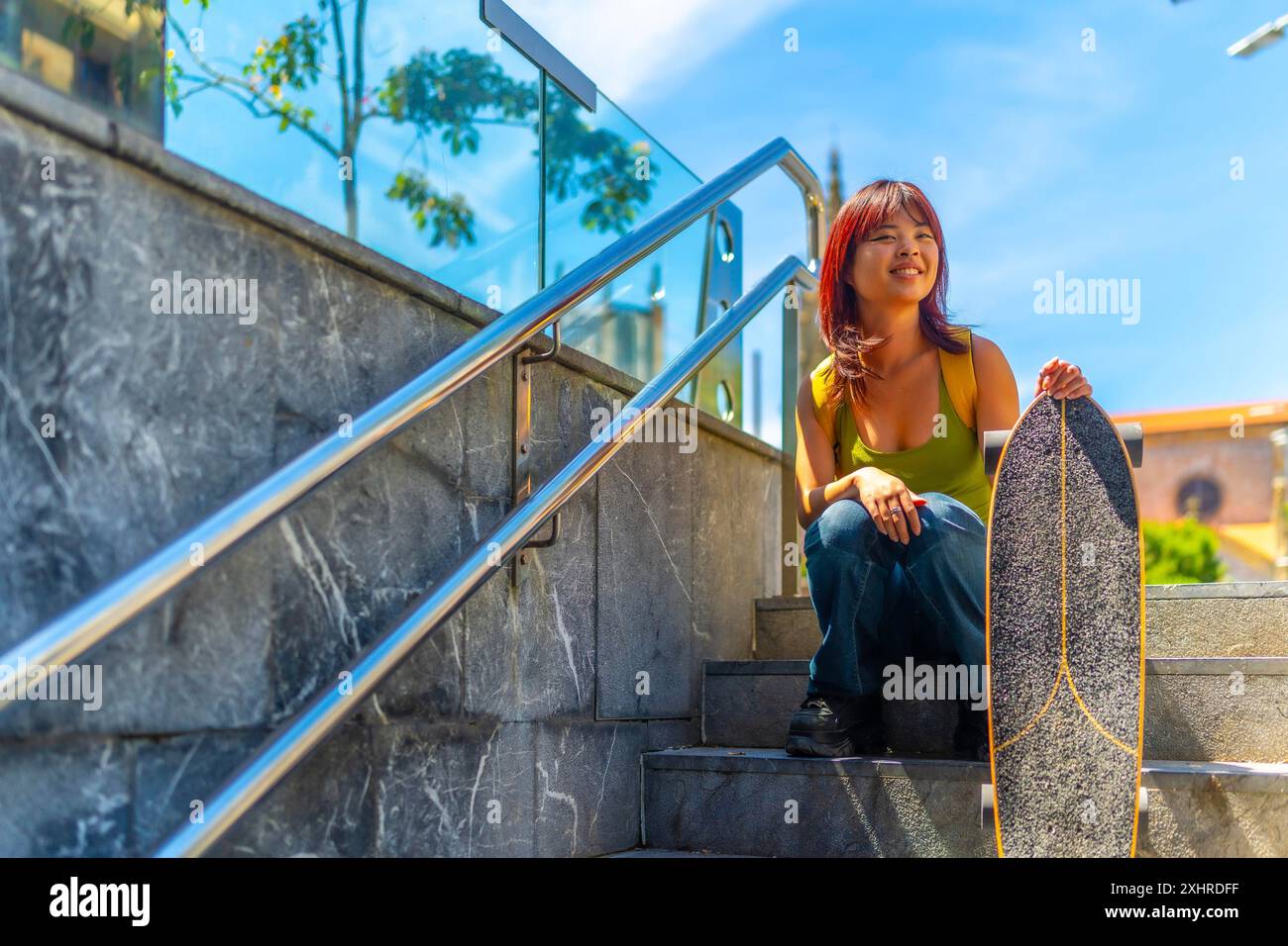 Coole junge asiatische Frau mit Skateboard, die im Sommer in der Stadt auf Treppen sitzt Stockfoto