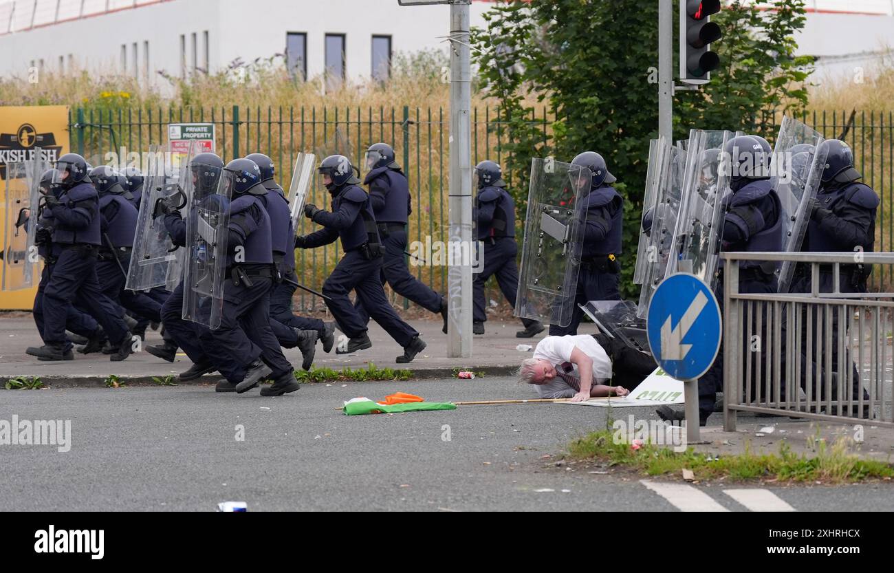 Ein Demonstrant mit einem Schild und einer Flagge wird auf den Boden gestoßen, als er von einer Linie von Beamten der Gardai-Einheit angegriffen wird, nachdem mehrere Brände auf dem ehemaligen Standort der Crown Pats Factory in Coolock, Nord-Dublin, ausgebrochen sind. Bilddatum: Montag, 15. Juli 2024. Stockfoto