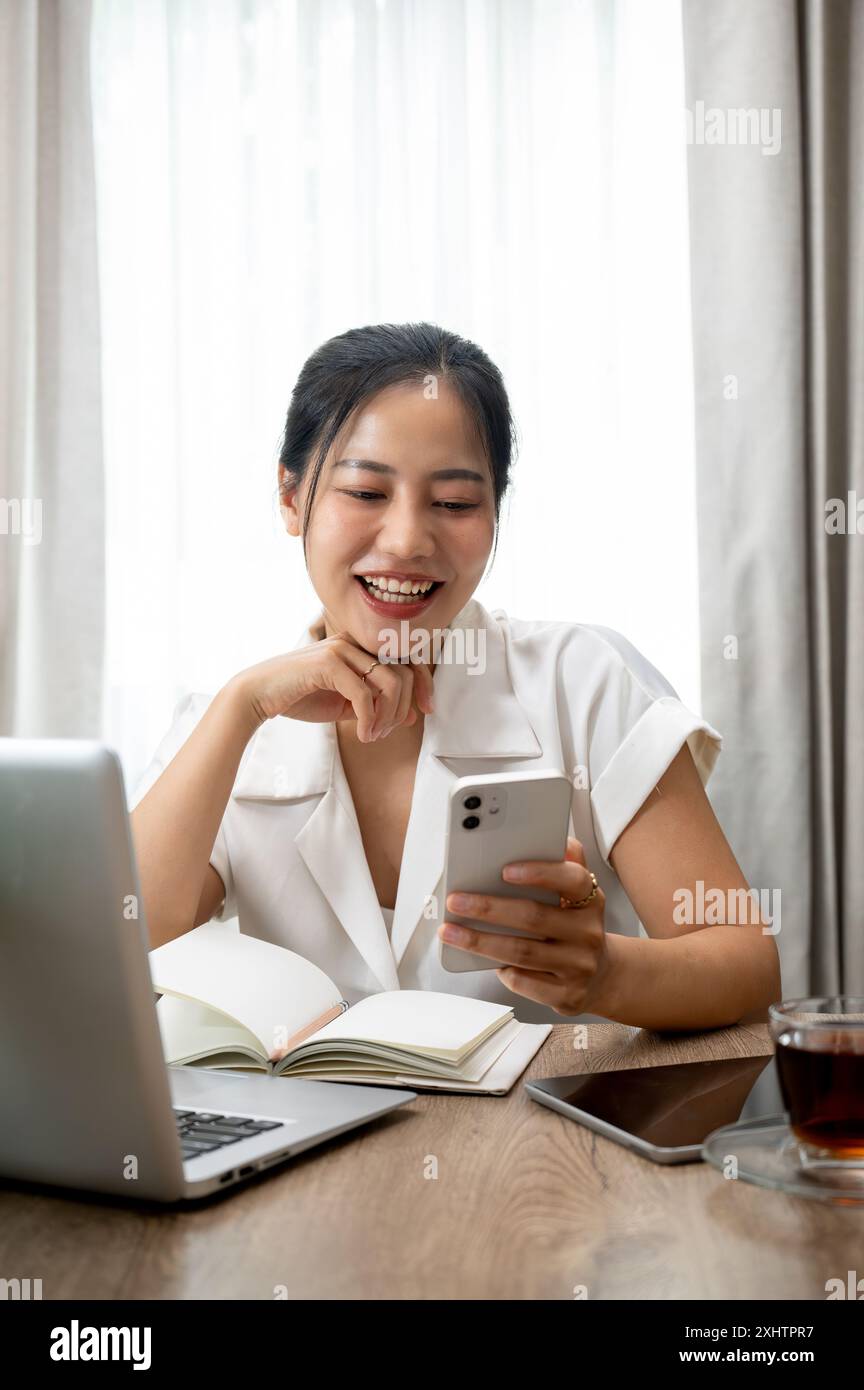 Eine attraktive asiatische Frau, die ihr Smartphone an ihrem Schreibtisch benutzt, Nachrichten liest, Chat beantwortet, Blogs im Internet liest. Menschen und Wireless-Technologie Stockfoto