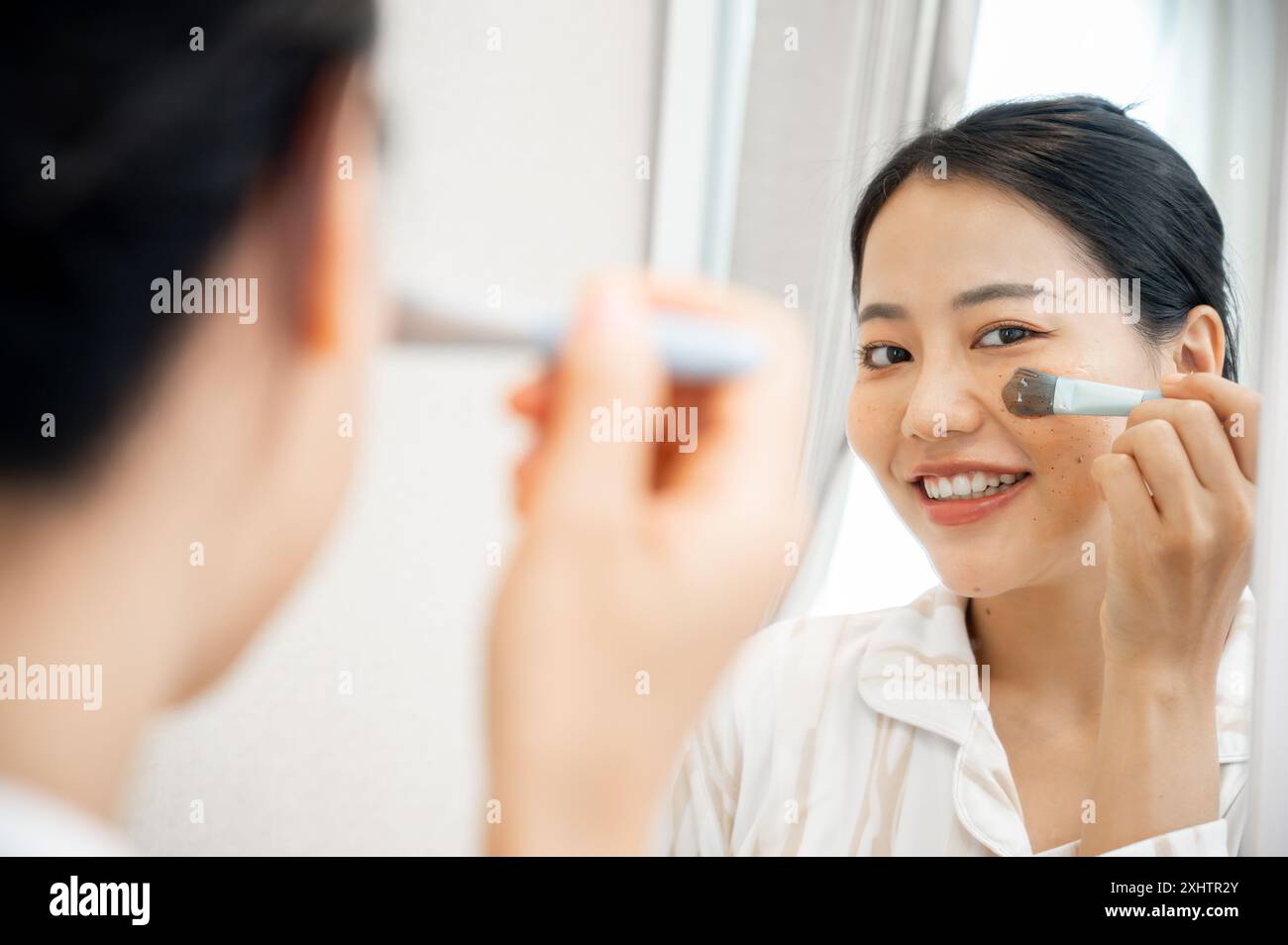 Eine schöne junge asiatische Frau im Pyjama trägt ihre Gesichtsmaske mit einem Pinsel vor dem Spiegel auf und macht ihre Selbstpflegeroutine bei Hom Stockfoto