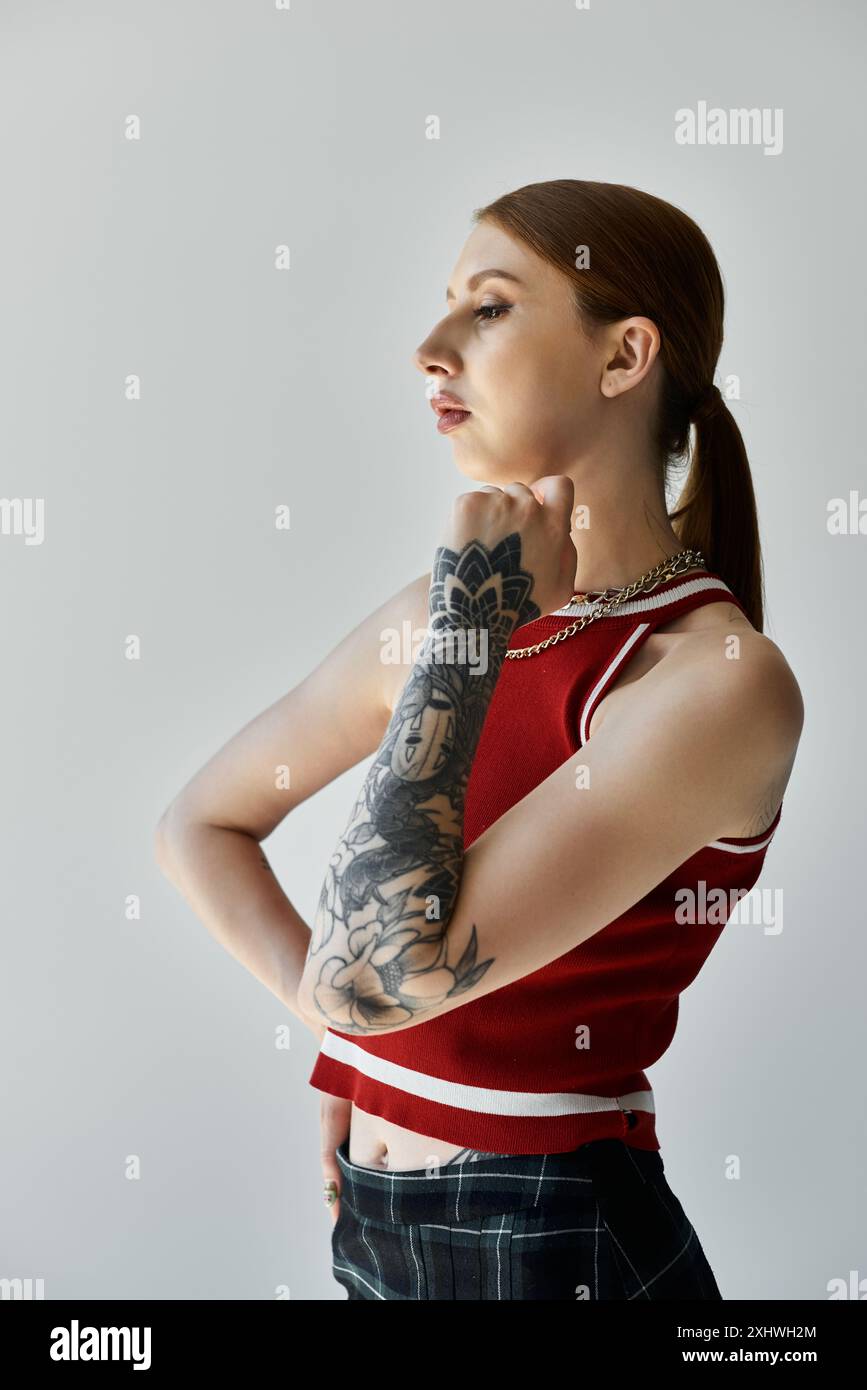 Eine junge Frau mit einem vollärmligen Tattoo steht vor einem grauen Hintergrund, trägt ein rotes Tanktop und einen karierten Rock. Stockfoto