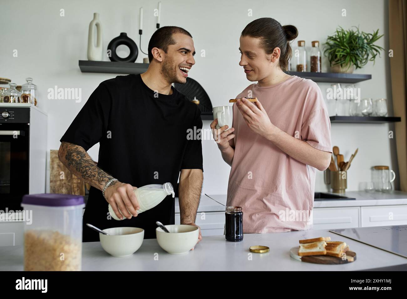 Zwei schwule Männer frühstücken gemeinsam in ihrer modernen Küche; einer gießt Milch in eine Schüssel, der andere lächelt mit Toast. Stockfoto