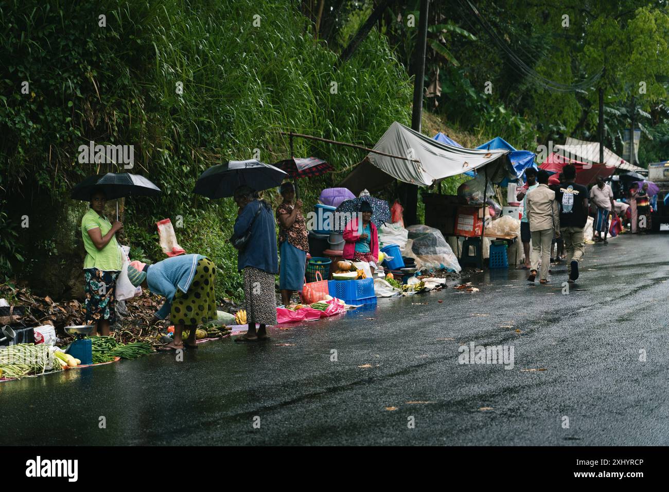 Regnerische Straßenmarktszene mit Leuten, die Regenschirme und Einkaufstaschen auf einer nassen Straße in Ella, Sri Lanka, tragen. Stockfoto