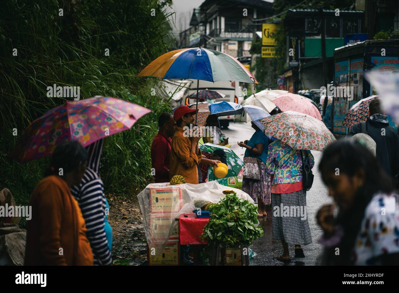 Regnerische Straßenmarktszene mit Leuten, die Regenschirme und Einkaufstaschen auf einer nassen Straße in Ella, Sri Lanka, tragen. Stockfoto