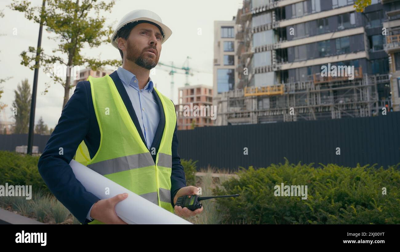 Kaukasischer Bauunternehmer Bauingenieur Elektroinstandhaltungsarbeiter, der einen Bauplan hält, männlich, der auf den Bauerbauer der Stadt blickt Stockfoto