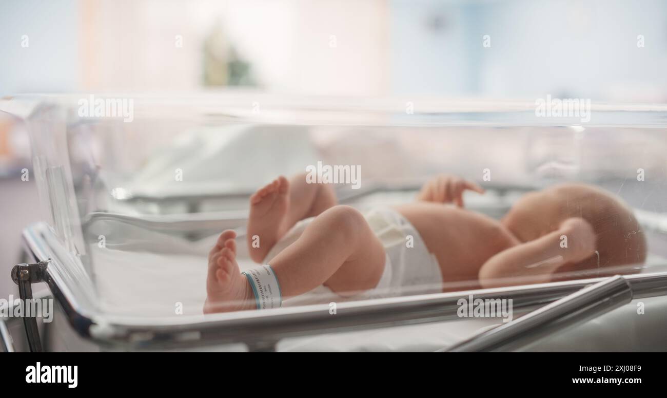 Niedliches kleines kaukasisches Neugeborenes, das im Bassinet in einem Entbindungskrankenhaus liegt. Porträt eines winzigen, verspielten und energiegeladenen Kindes mit einem Namensschild auf dem Bein. Gesundheit, Schwangerschaft und Mutterschaft Konzept Stockfoto