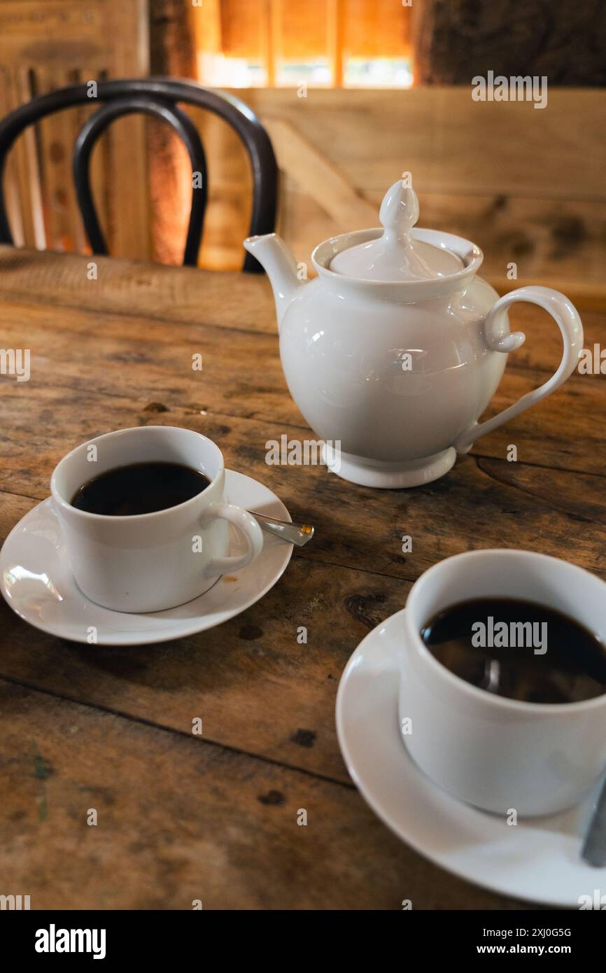 Ein Holztisch mit zwei Tassen Kaffee und einer weißen Teekanne. Die Umgebung befindet sich in einem kleinen ländlichen Restaurant und die Sonne erleuchtet die Kulisse. Stockfoto