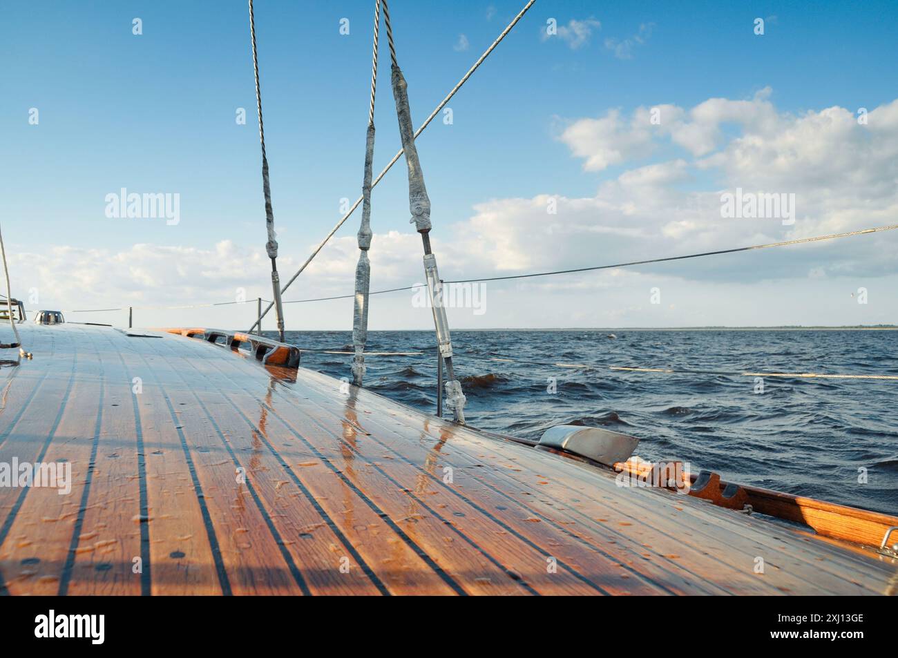 Segeln in einem Meer bei sonnigem Wetter. Himmel teilweise bedeckt mit Wolken, kleine Wellen auf der Oberfläche. Glänzendes hölzernes Yachtdeck bedeckt mit Wassertropfen. Stockfoto