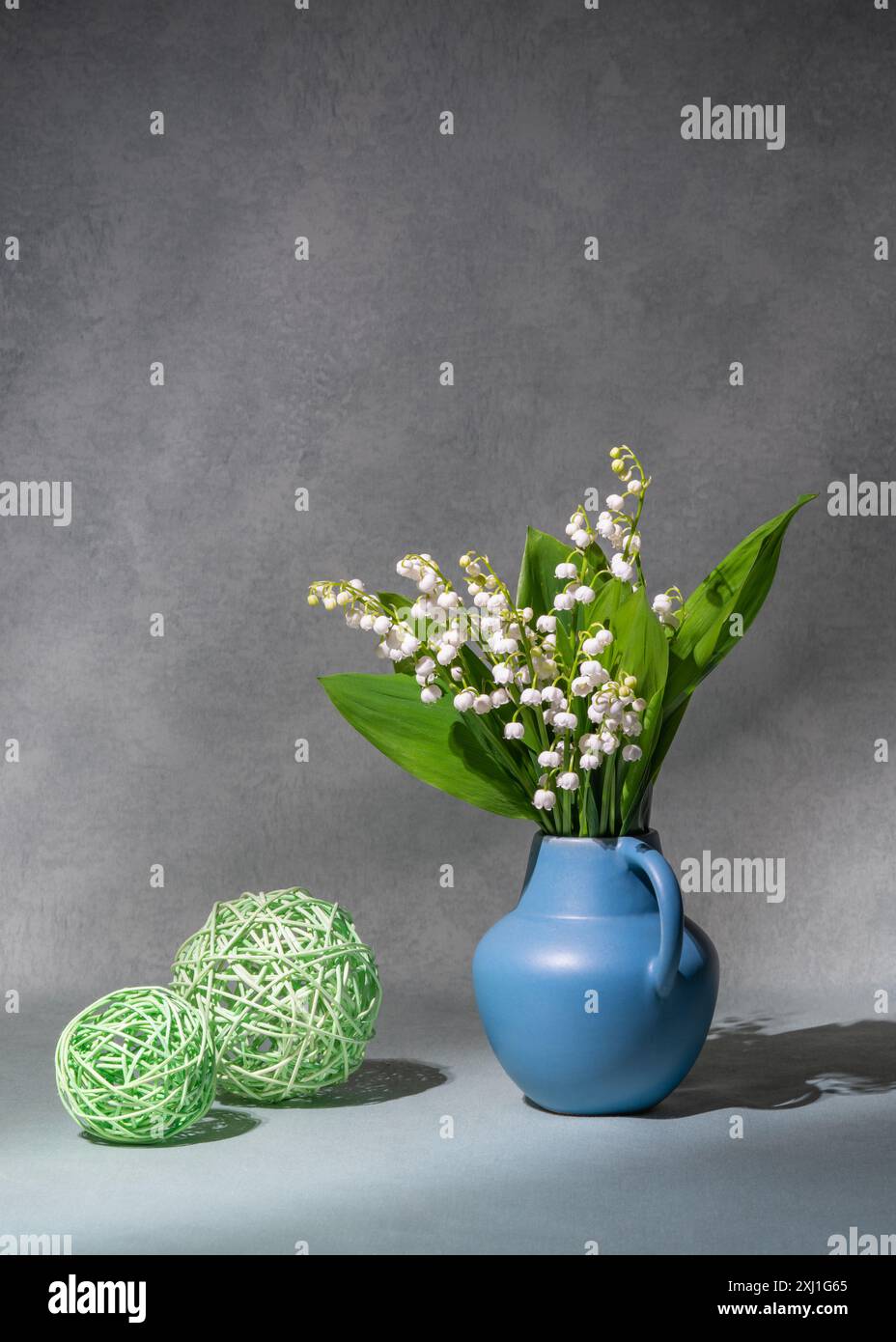 Ein Blumenstrauß aus Frühlingsblumen, Maiglöckchen aus Tal in einer blauen Vase, auf grauem Hintergrund. Es gibt zwei grüne Bälle in der Nähe. Stillleben, Postkarte, Kopie Spac Stockfoto