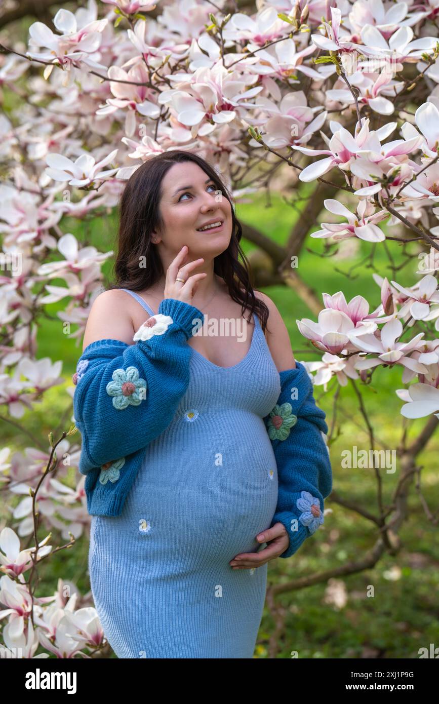 Porträt einer niedlichen schwangeren Frau im Park, gekleidet in einem blauen Kleid zwischen Magnoli-Blumen. Die werdende Mutter schaut zärtlich auf einen blühenden Baum. Stockfoto