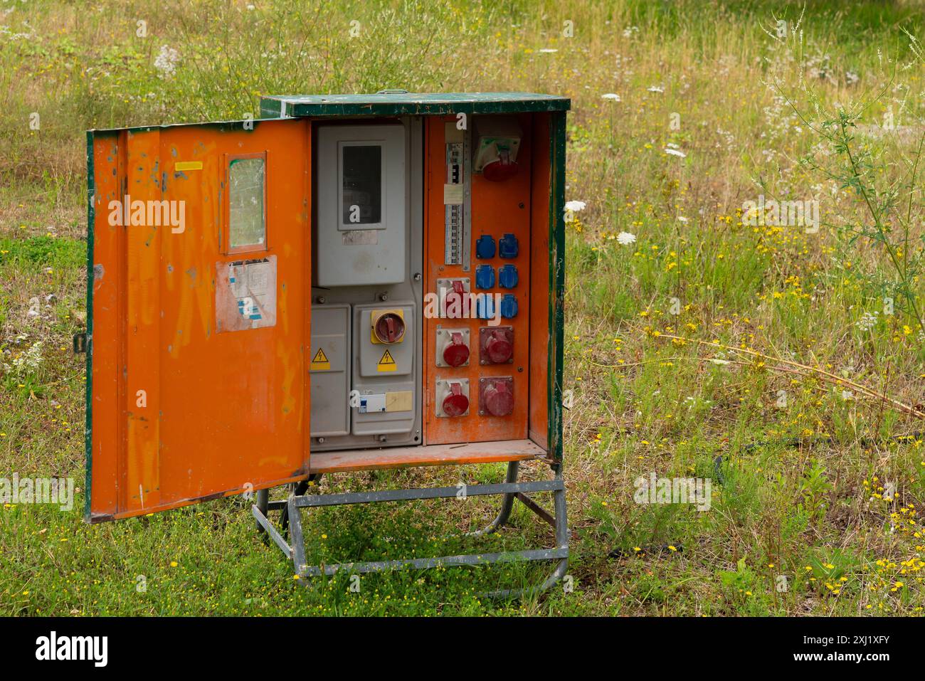 Ein alter orangefarbener Schaltkasten mit offener Metalltür, in dem eine Vielzahl von Steckdosen, Schaltern und ein Messgerät zu sehen sind. Die Box befindet sich auf einem Feld mit grünem Gras und gelben Wildblumen. Stockfoto