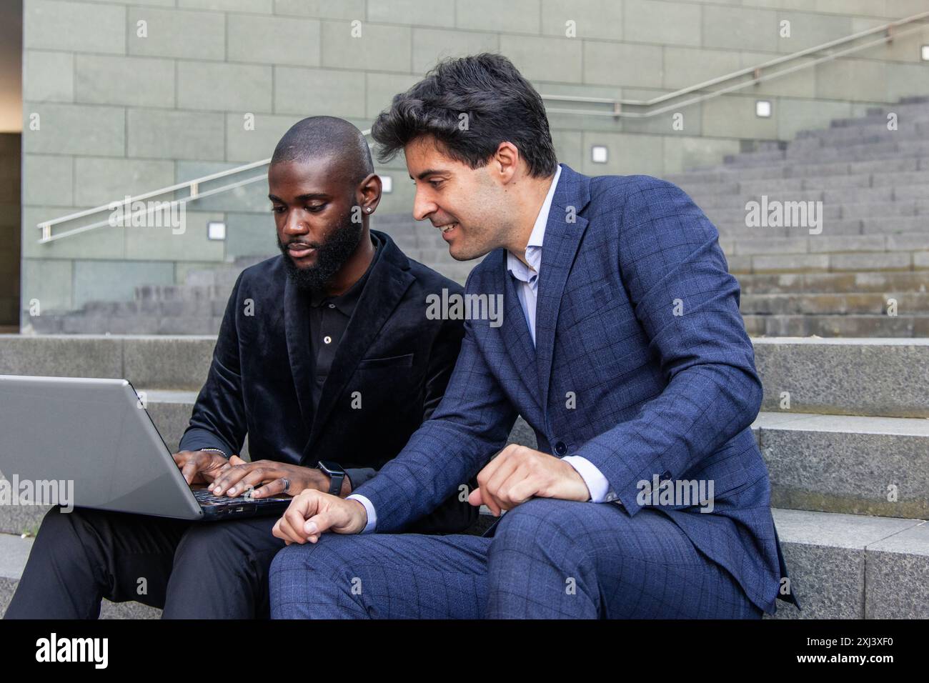 In einem Finanzviertel der Stadt arbeitet ein multikulturelles Duo von Geschäftsleuten, ein schwarzer Afrikaner und der andere Hispanic, an einem Laptop zusammen, während sie auf einem Platz sitzen Stockfoto