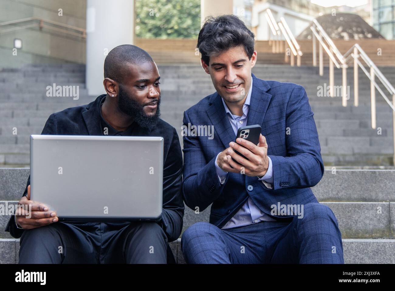 Ein hispanischer Geschäftsmann zeigt seinem afrikanischen Kollegen, der einen Laptop in der Hand hat, Unternehmensdaten auf dem Smartphone an, während er draußen auf einer Bürotreppe arbeitet.highl Stockfoto