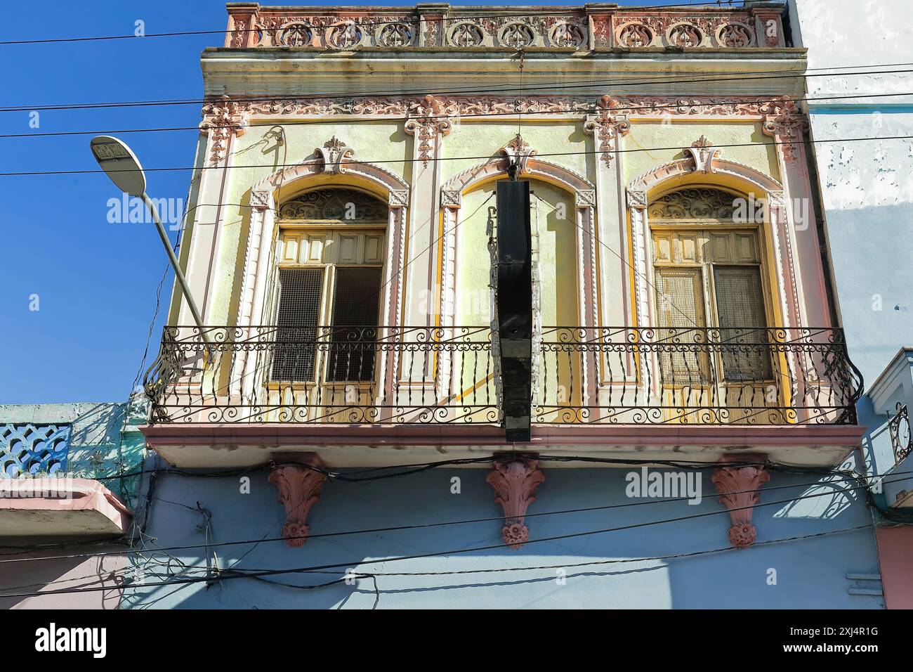 Fassade des eklektischen Gebäudes aus dem Jahr 502 mit blassgelb lackiertem ersten Stock und rosafarbenen Zierleisten, Balkon mit Eisengeländer auf geblümten Konsolen. Santiago-Kuba. Stockfoto