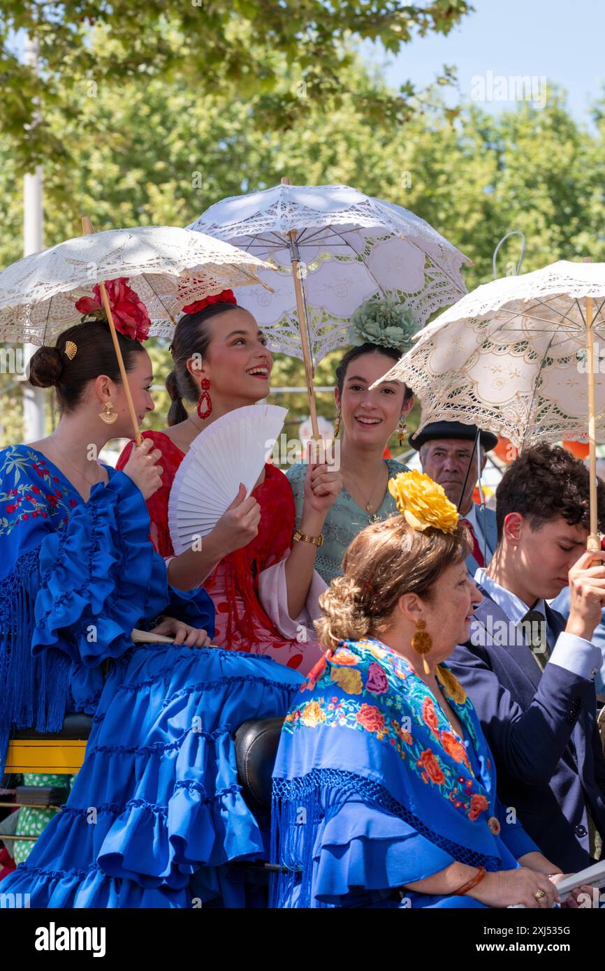 Frauen in leuchtenden Kleidern und Sonnenschirmen lächeln und halten Fans unter der Sonne, Mai-Festivals in Sevilla in Andalusien in Spanien Stockfoto